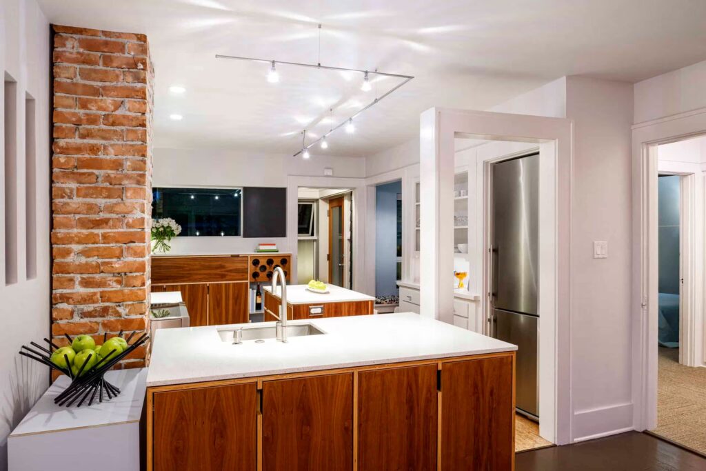 A cozy kitchen featuring a rustic brick wall, complete with modern cabinetry, a countertop, sink, and tap. The interior design includes drawers and cupboards, with an island in the center of the space. Bright natural light streams in through a window, illuminating the warm wooden floor and stylish furniture.
