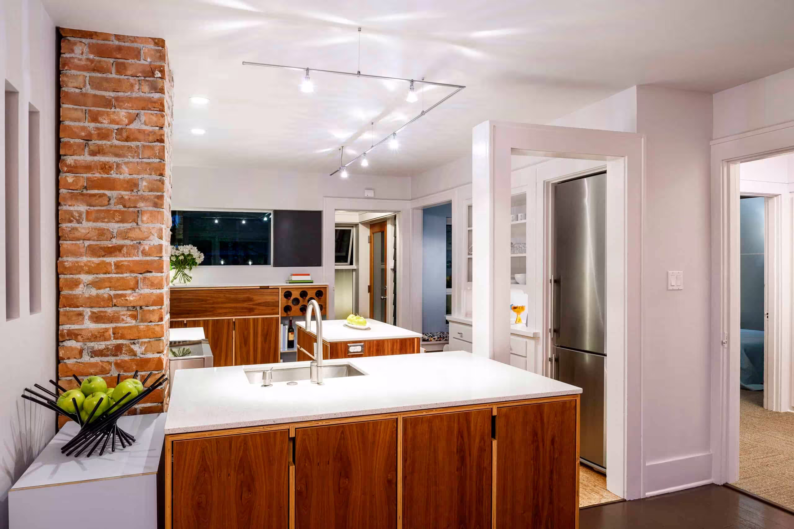 A cozy kitchen featuring a rustic brick wall, complete with modern cabinetry, a countertop, sink, and tap. The interior design includes drawers and cupboards, with an island in the center of the space. Bright natural light streams in through a window, illuminating the warm wooden floor and stylish furniture.