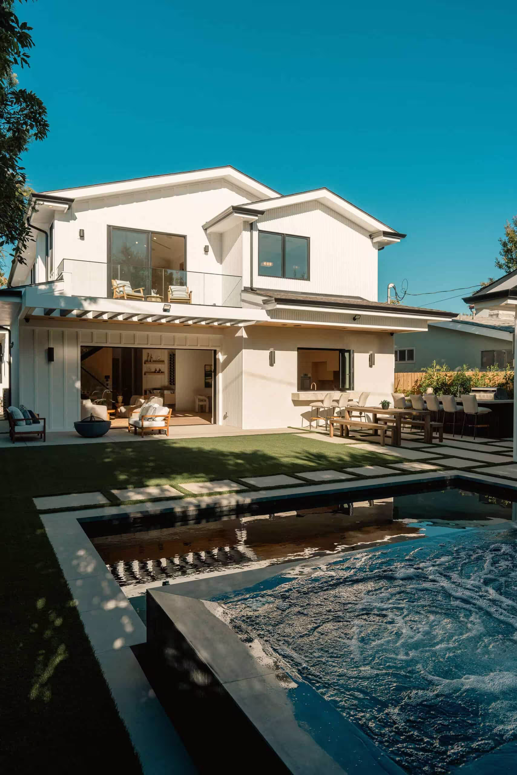 A house with a swimming pool in the front yard, surrounded by trees and under a clear sky. The property features windows reflecting the outdoor scenery.